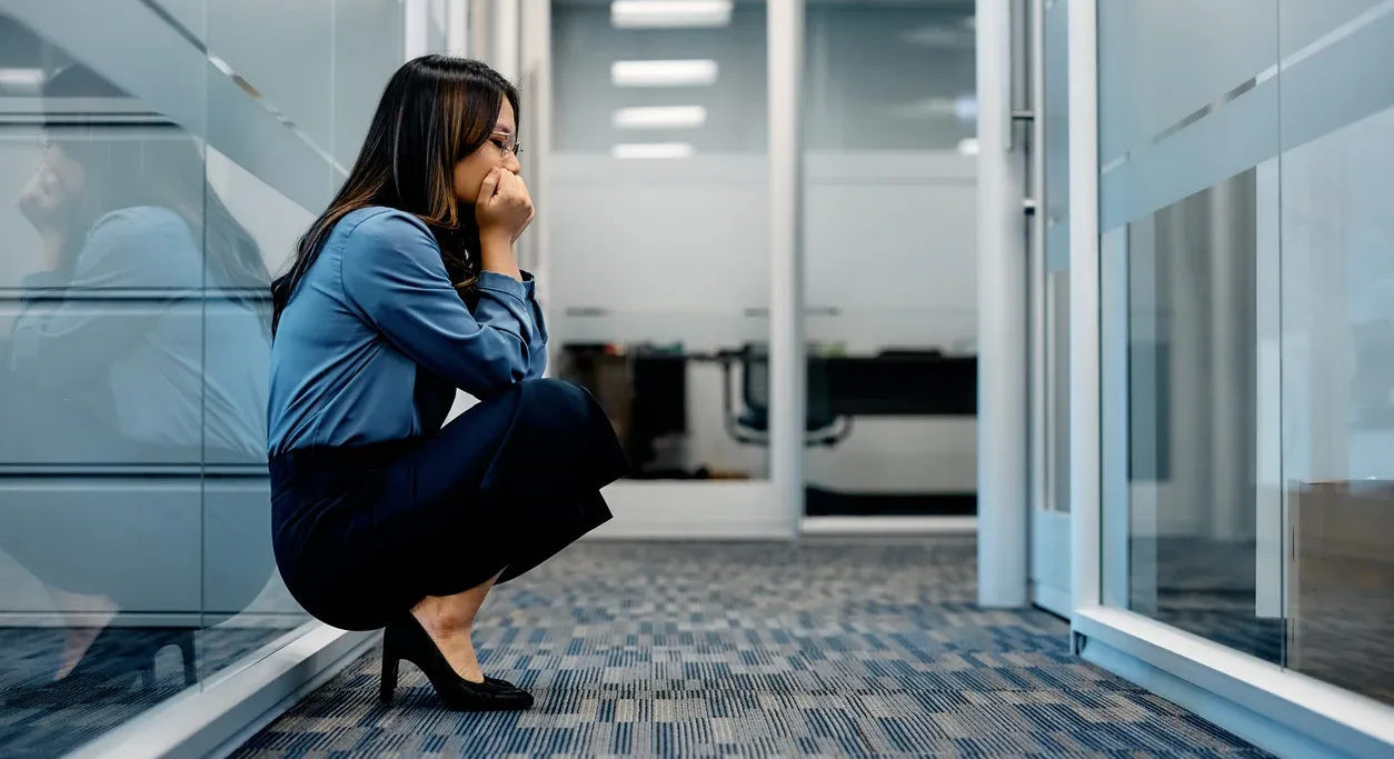 Woman in office hallway, squatting with hands on face, looking distressed.
