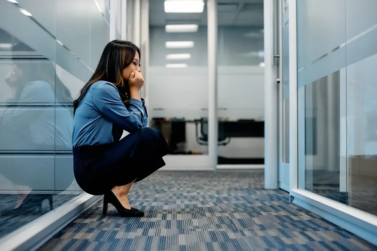 Woman in office hallway, squatting with hands on face, looking distressed.
