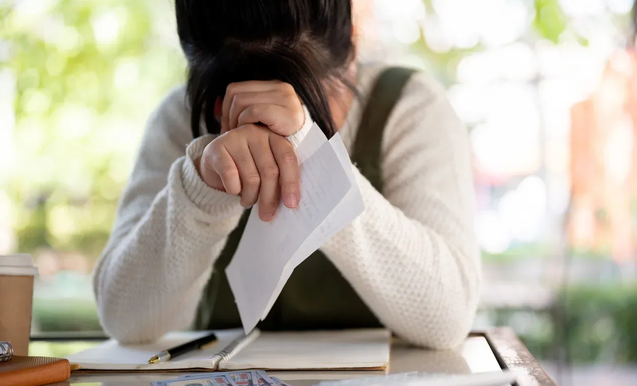 Woman sitting at a table with head in hands, holding a paper, appearing distressed; notebook and coffee visible.