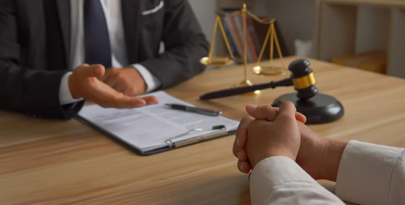 A person in suit gesturing to a client at a desk; scales of justice and gavel in background.