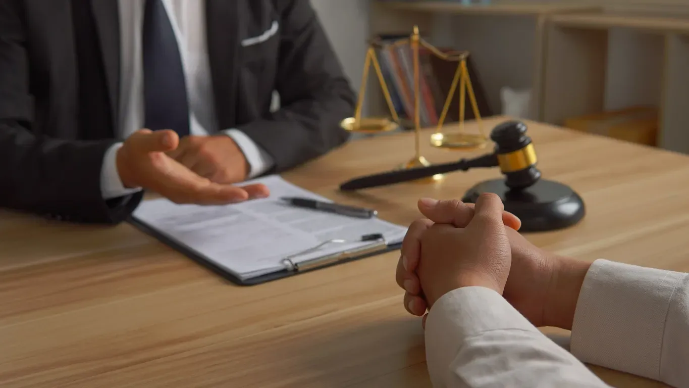 A person in suit gesturing to a client at a desk; scales of justice and gavel in background.