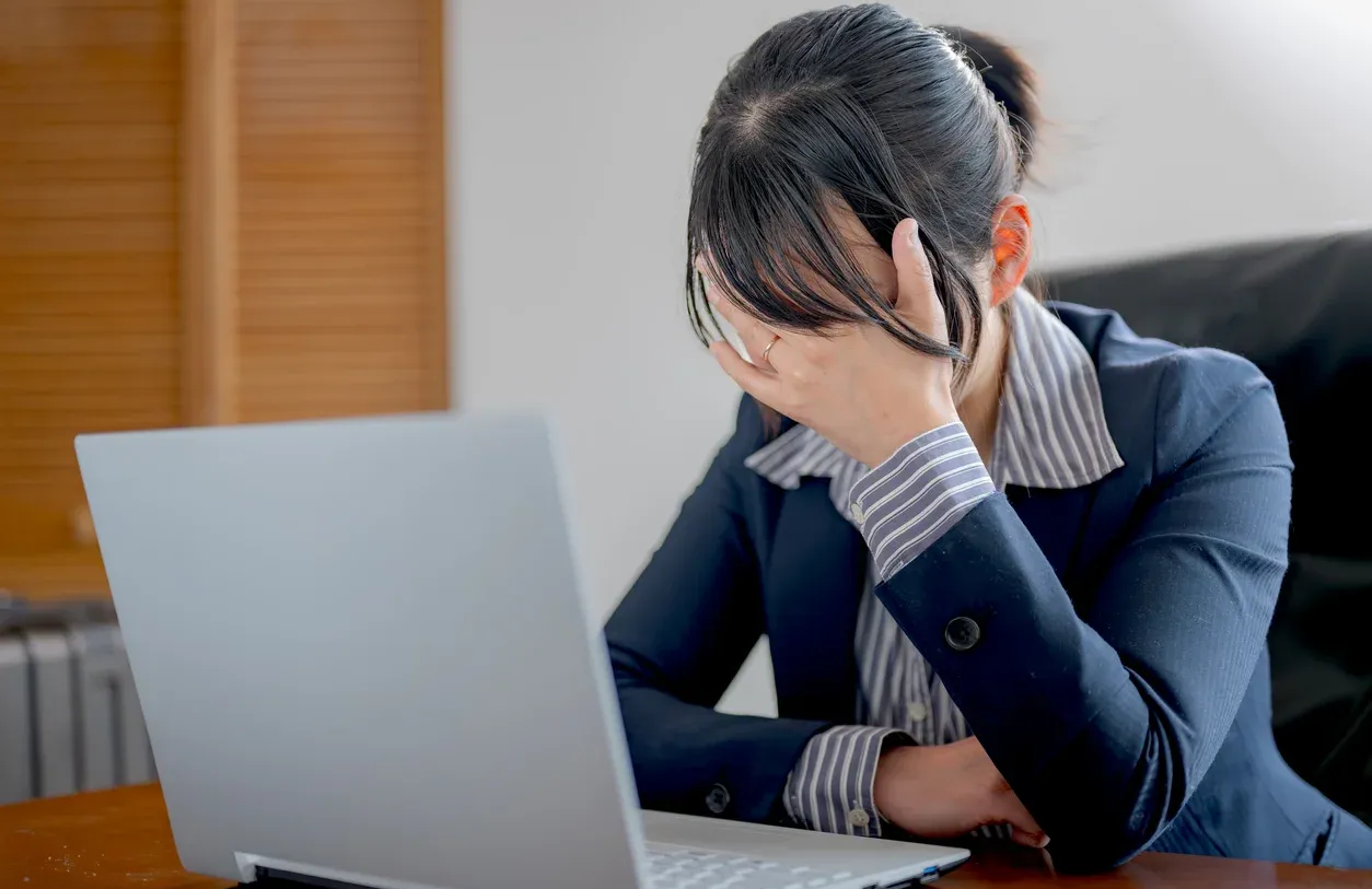 Woman in blue blazer, face in hands, sits at desk with laptop, appearing stressed.