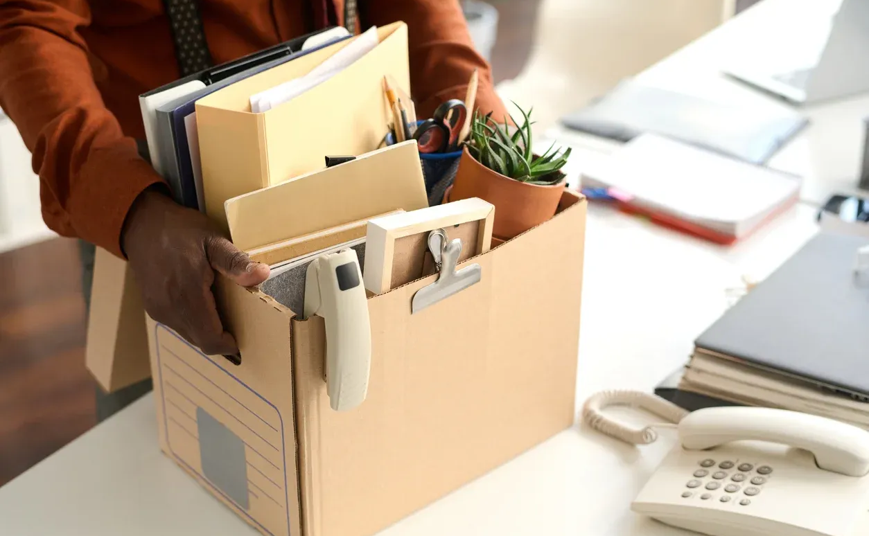 Person holding a cardboard box filled with office items, packing up at a desk.