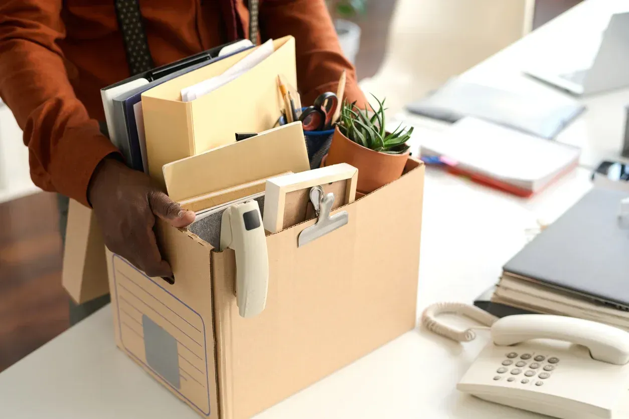 Person holding a cardboard box filled with office items, packing up at a desk.