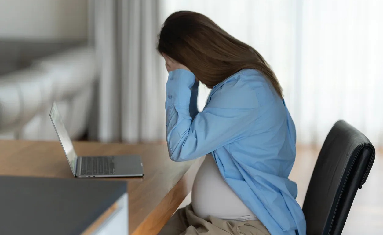 Pregnant woman, stressed, holding her face at a desk with a laptop.