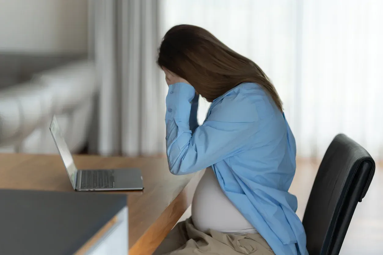 Pregnant woman, stressed, holding her face at a desk with a laptop.