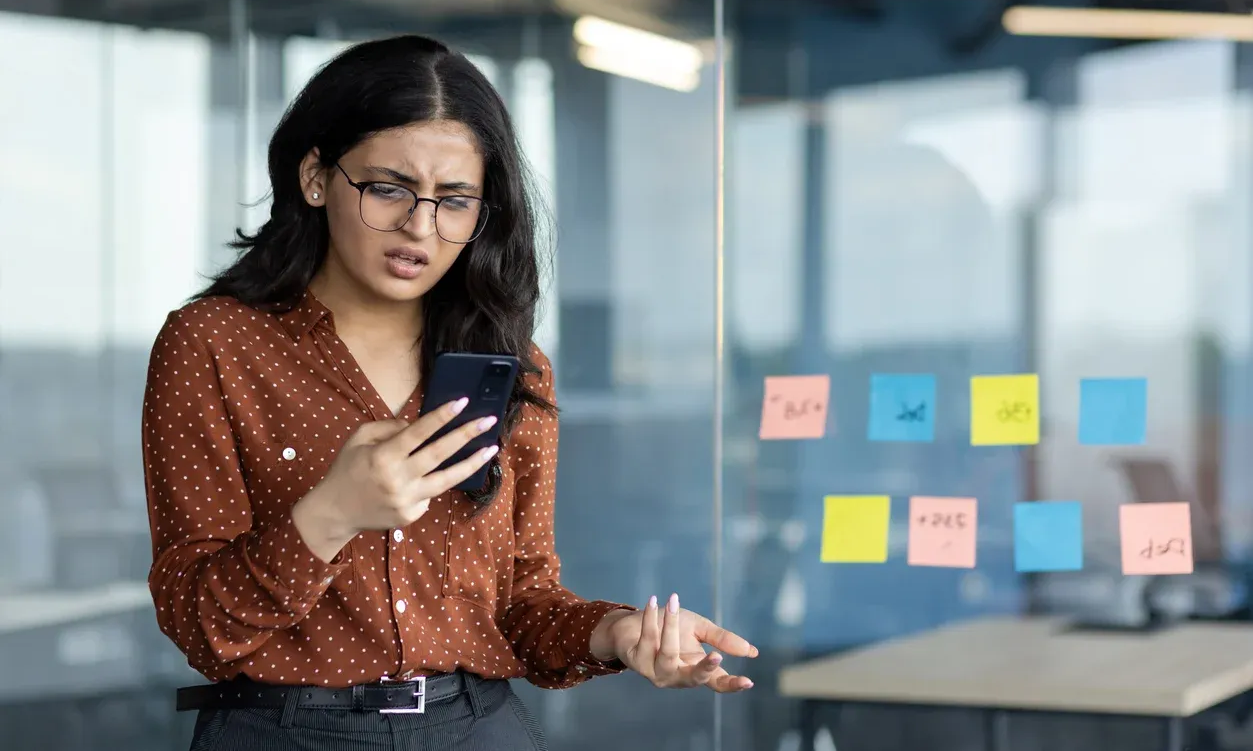 Woman in glasses looks confused and upset at her phone in an office with sticky notes on the glass.
