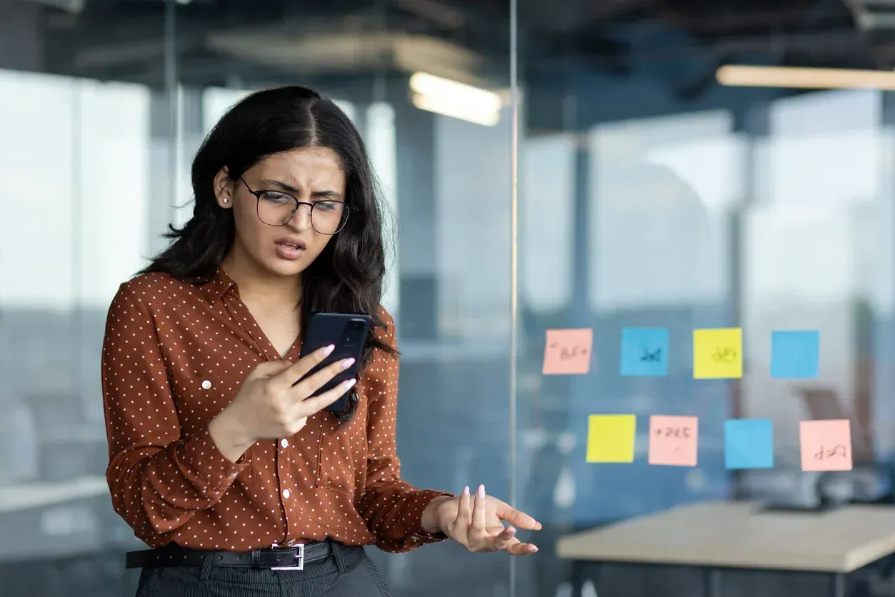 Woman in glasses looks confused and upset at her phone in an office with sticky notes on the glass.