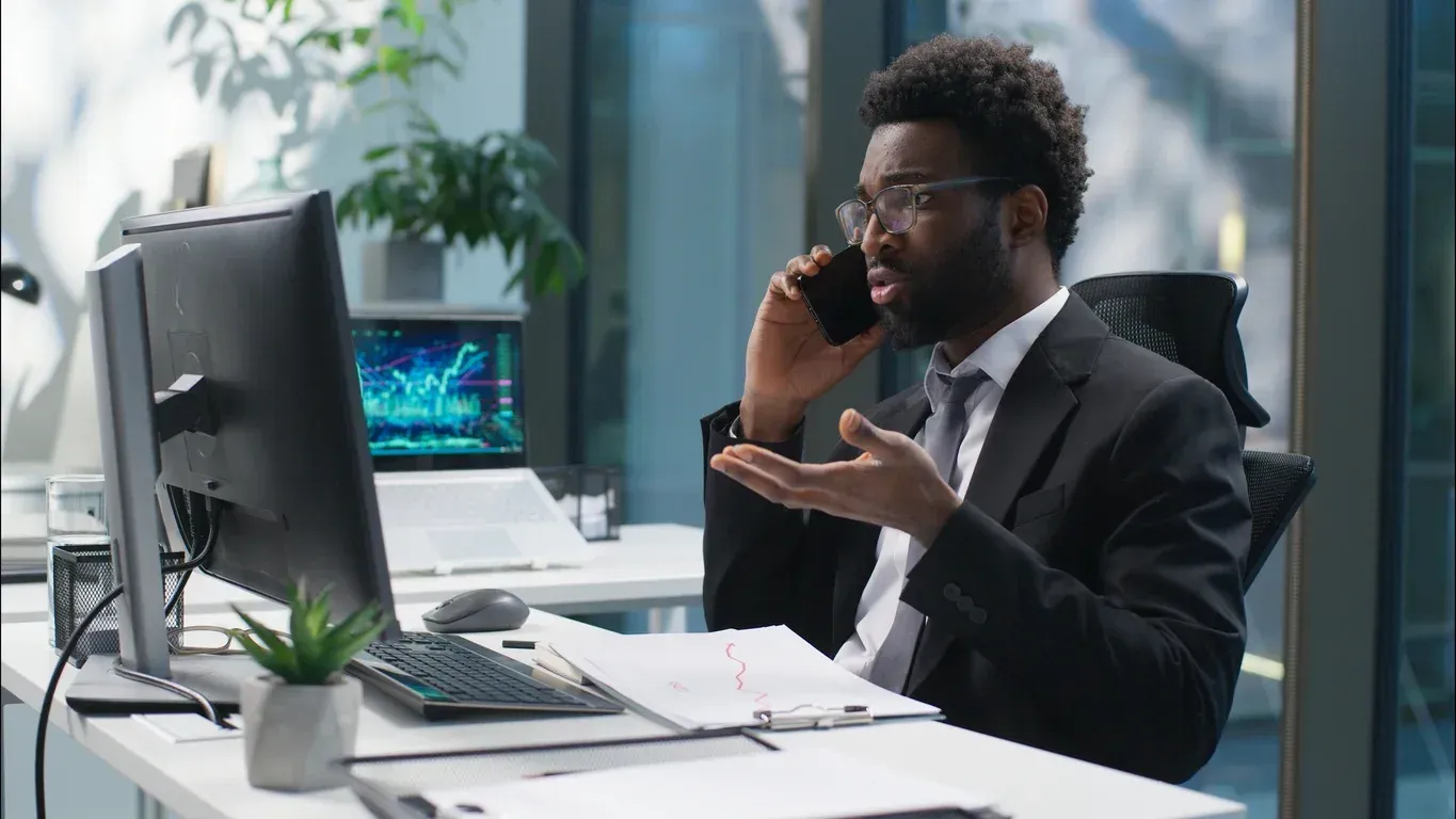 Man in suit talking on phone at a desk with computer, charts visible, gesturing, indoor office setting.