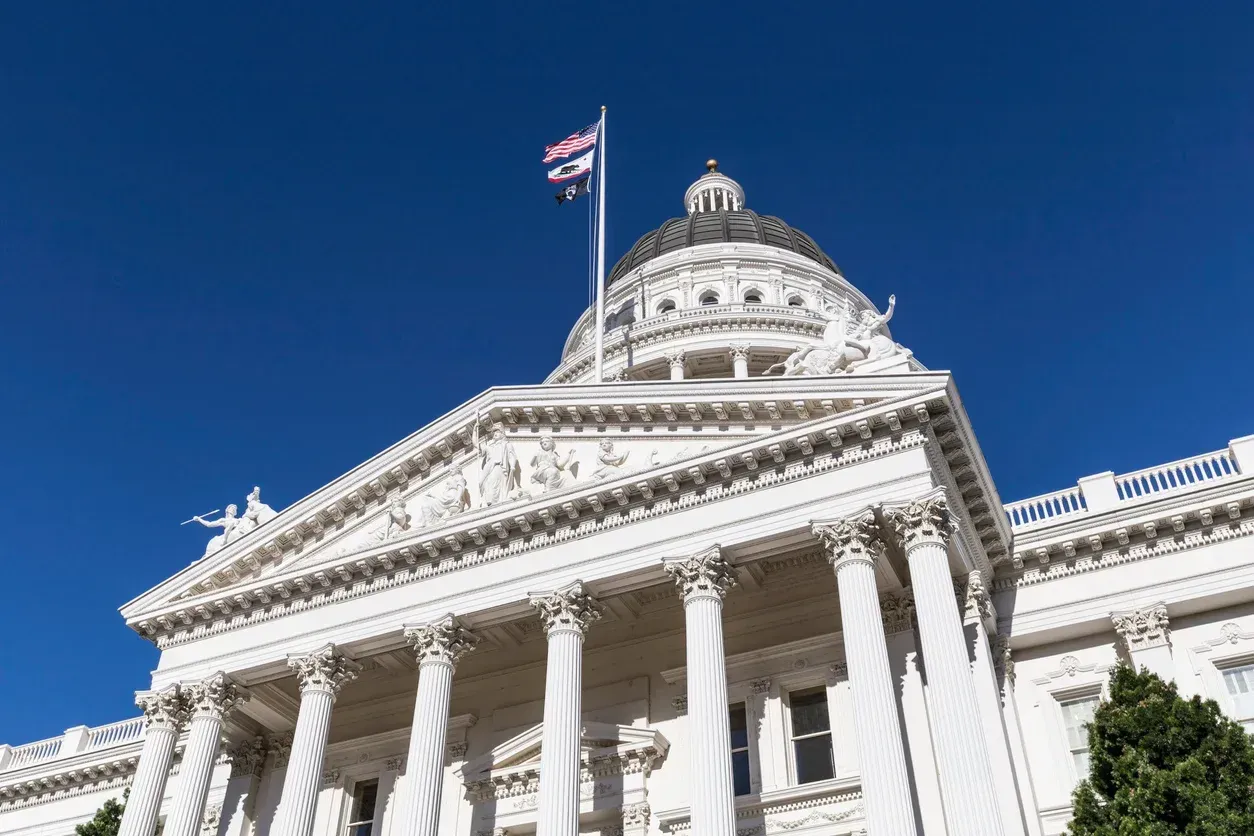 California State Capitol building with white columns, dome, and flag against a blue sky.
