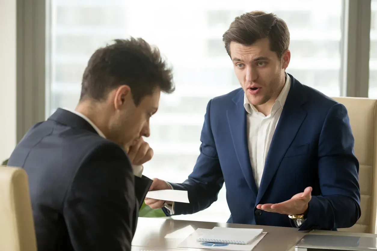 Man in suit yelling at another man at a table, holding a paper; office setting.