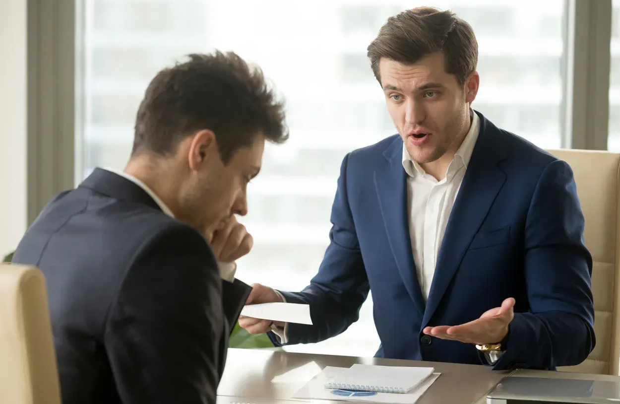 Man in blue suit gestures angrily at a seated man, holding papers. Office setting.