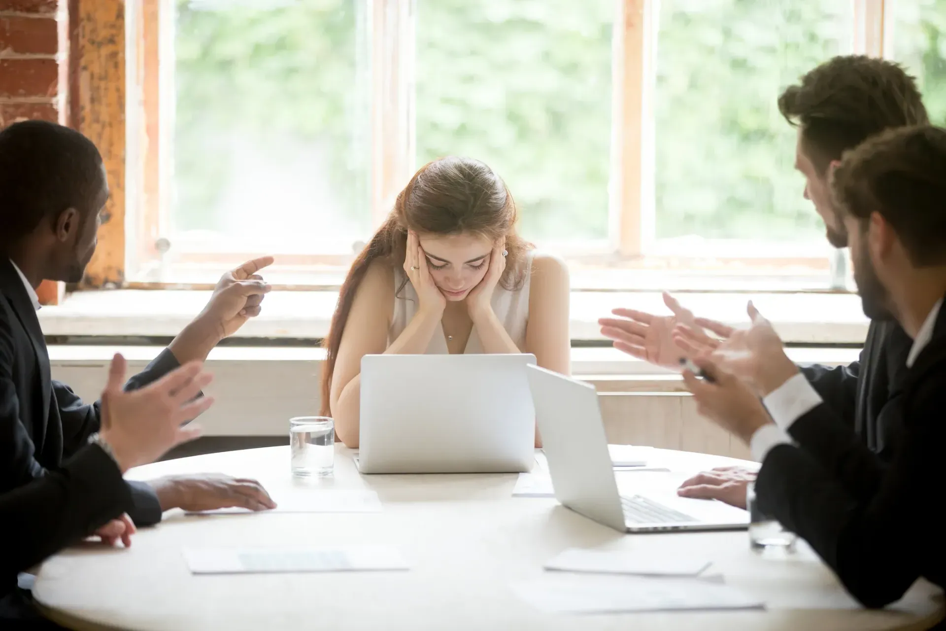 Woman looks stressed at a table, surrounded by men pointing and gesturing in an office setting.