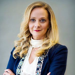 A woman in a blue jacket and white shirt is standing with her arms crossed.
