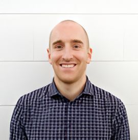 A man in a plaid shirt is smiling in front of a white wall.