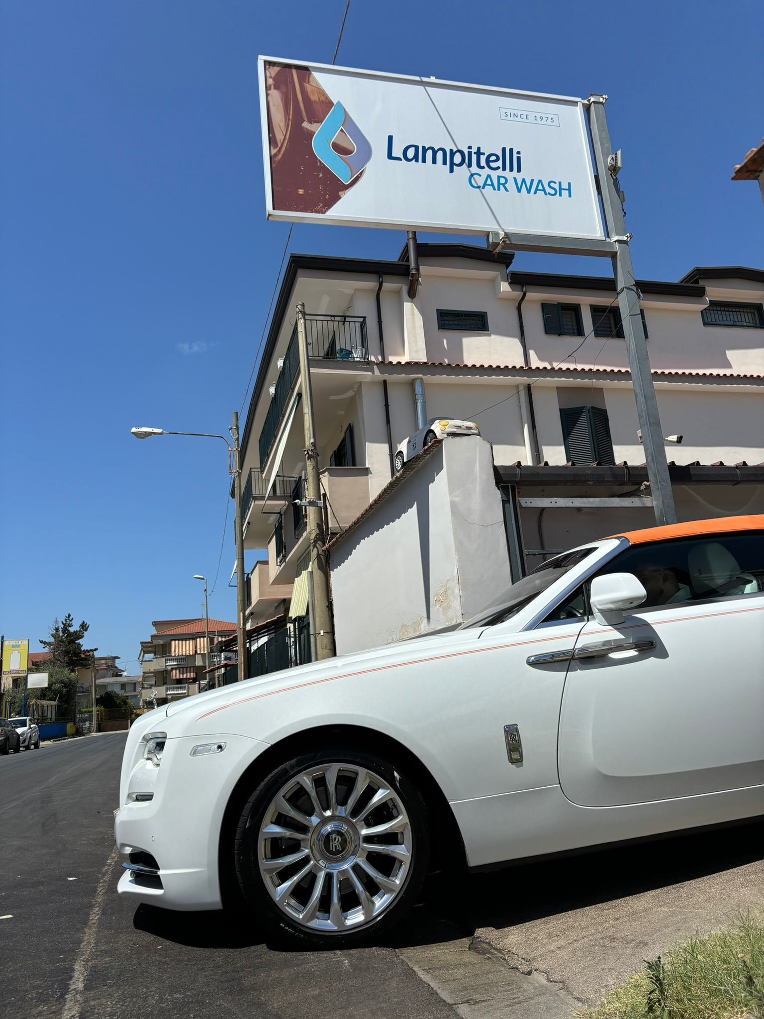 Una Rolls Royce bianca parcheggiata sulla strada davanti all'insegna di una farmacia, sotto un cielo azzurro.