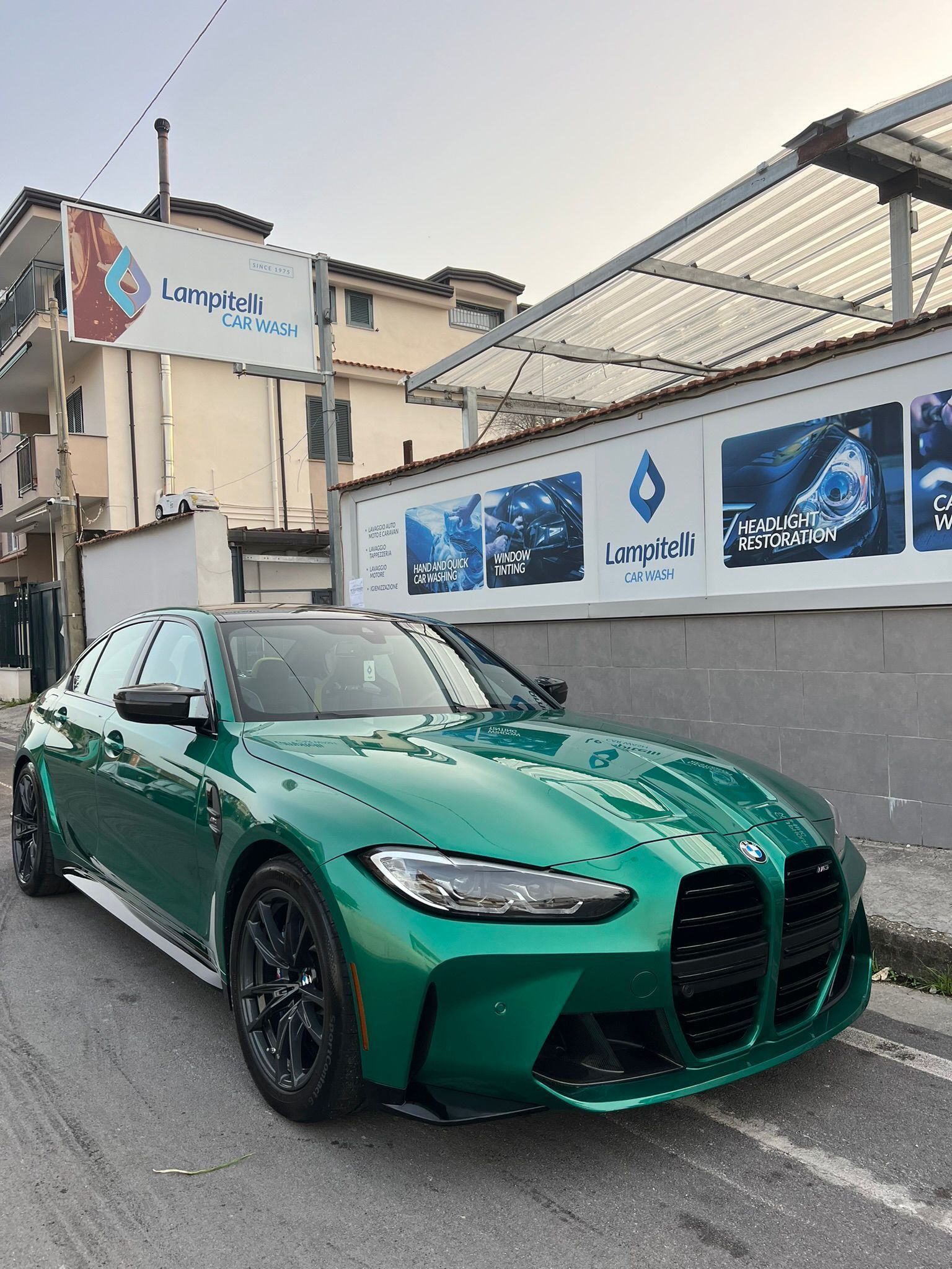 Una BMW M4 verde parcheggiata davanti a un autolavaggio con un edificio, un cartello e pannelli solari sullo sfondo.