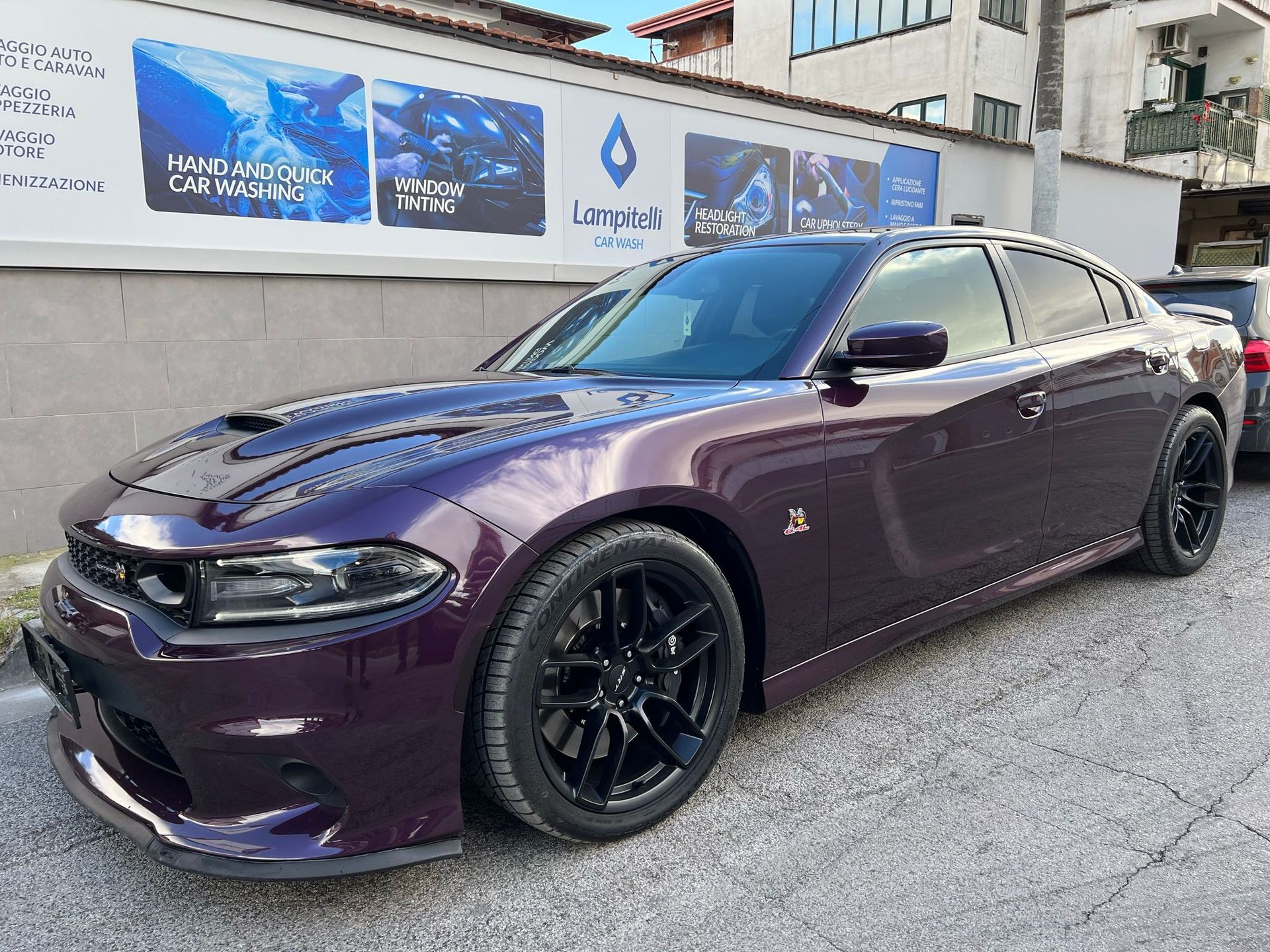 Una Dodge Charger viola parcheggiata davanti a un edificio con cartelloni pubblicitari. Cerchi neri e presa d'aria sul cofano.