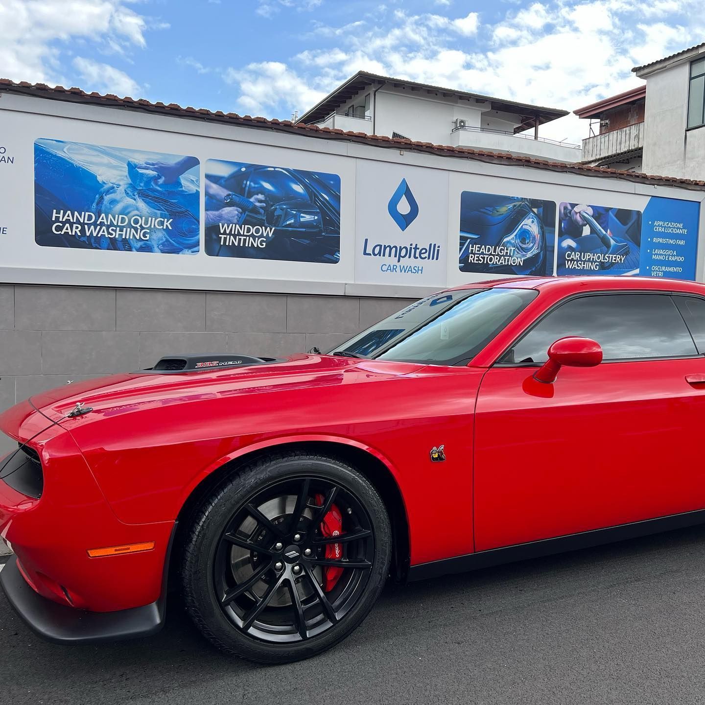 Una Dodge Challenger rossa parcheggiata davanti a un autolavaggio con striscioni pubblicitari in una giornata di sole.