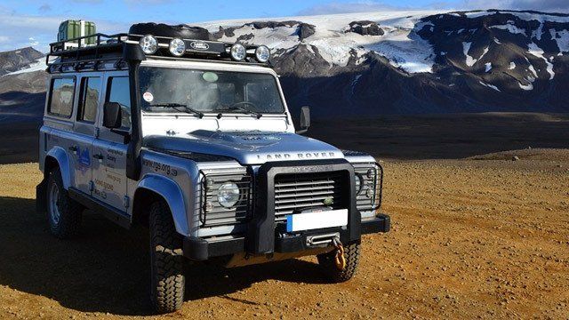 A land rover defender is parked in a dirt field with mountains in the background.