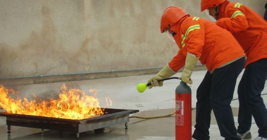 I vigili del fuoco, con indosso l'equipaggiamento protettivo arancione, spengono un incendio in una vasca di metallo con un estintore.
