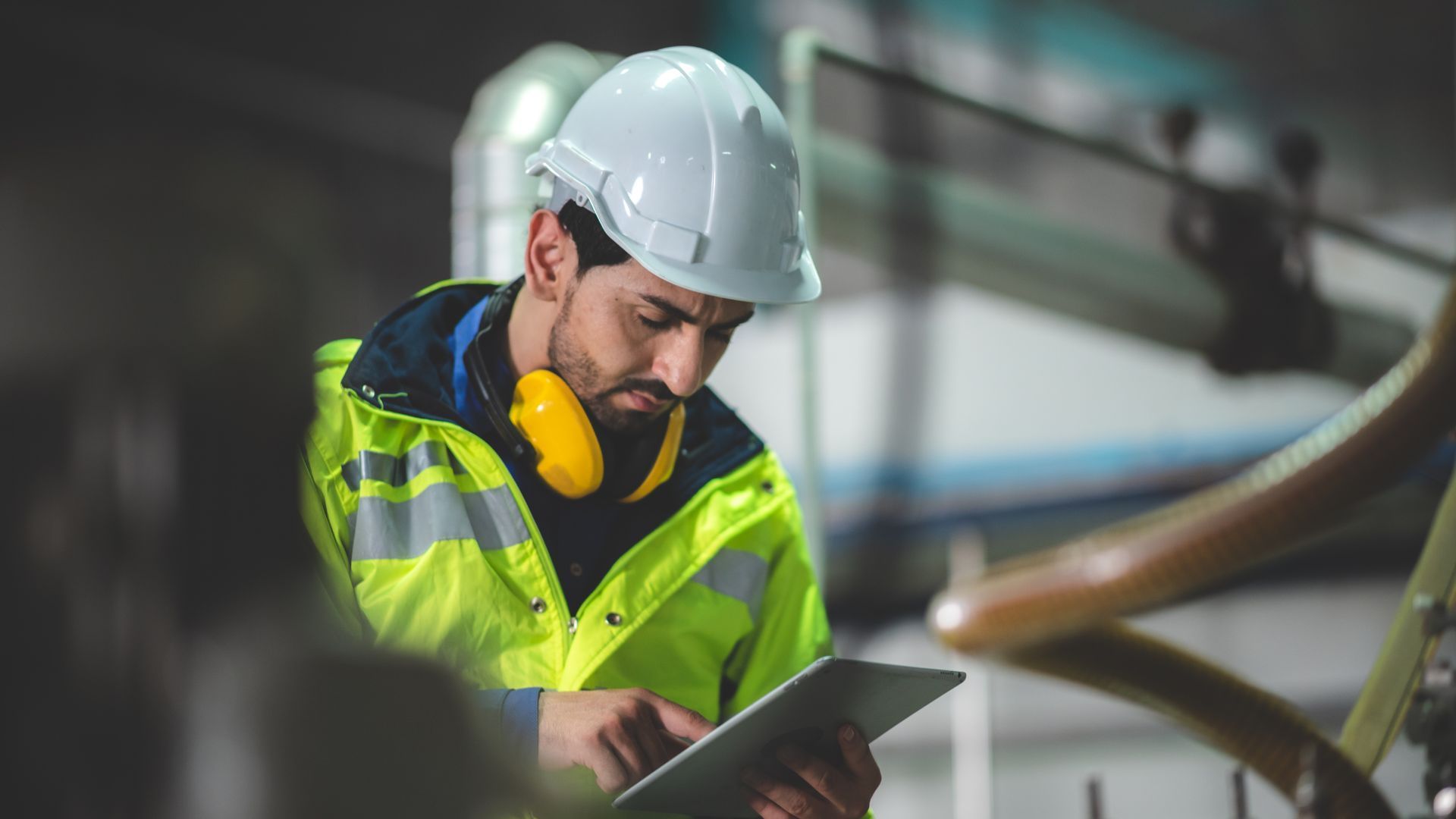 Uomo con equipaggiamento di sicurezza che guarda un tablet in un ambiente di fabbrica.