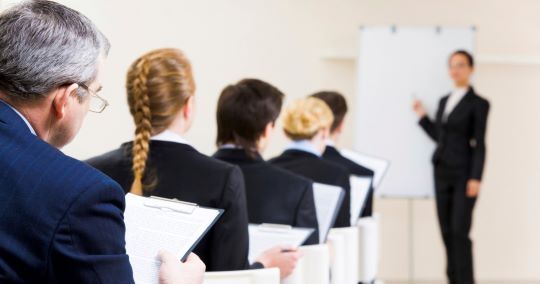 Una donna in giacca e cravatta si presenta al pubblico in una sala conferenze dalle pareti bianche, indicando una lavagna.