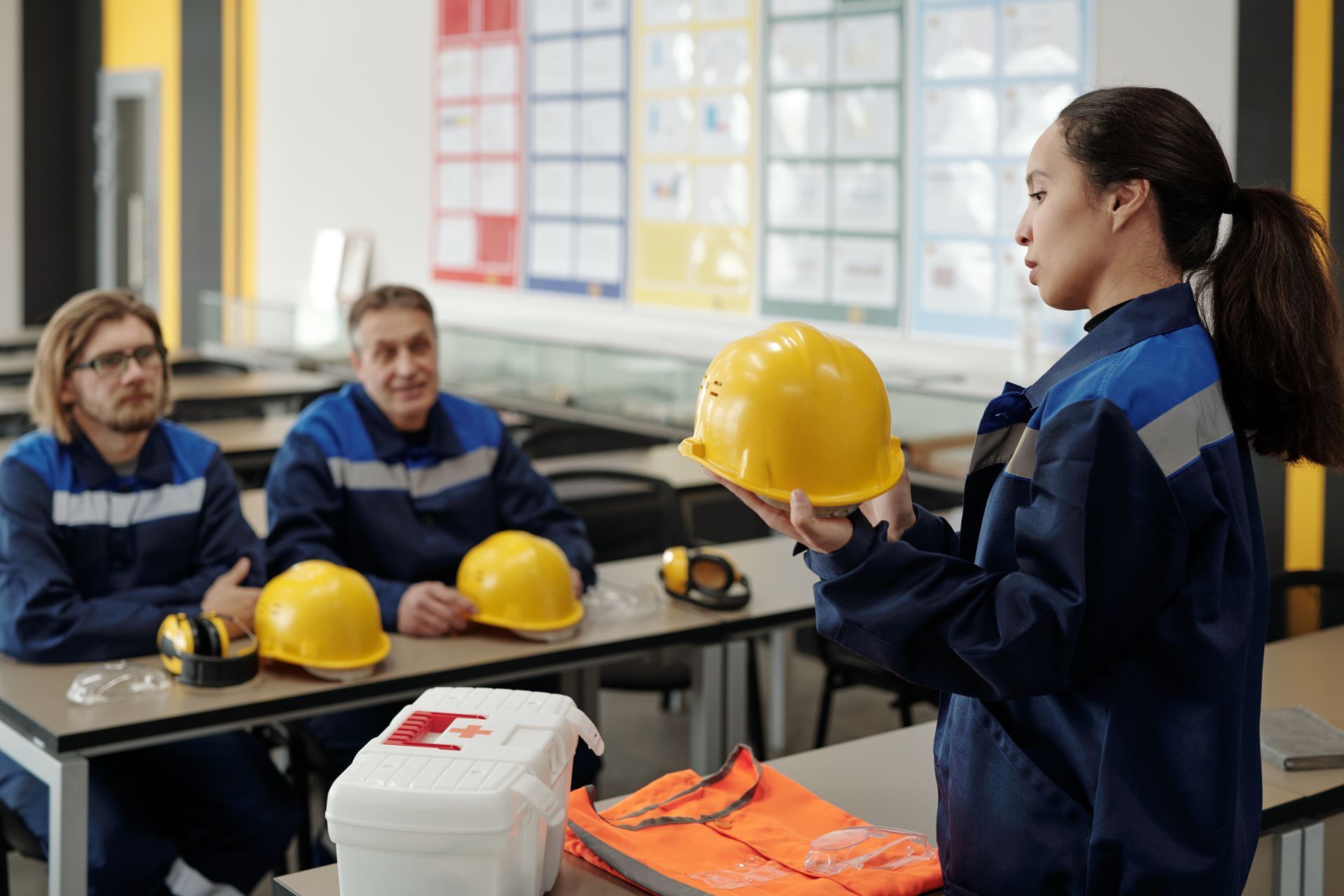 Una donna in tuta blu mostra il casco di sicurezza a due colleghi seduti. Ambiente scolastico.