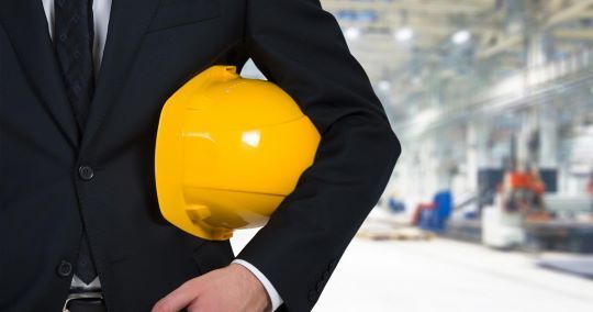 Persona in tuta che tiene in mano un casco giallo in un ambiente industriale.