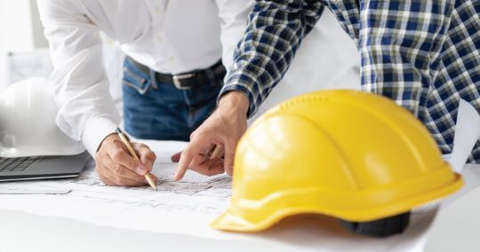 Due persone esaminano i progetti, con un casco giallo in primo piano.