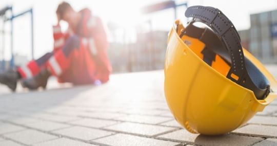 Casco giallo sul marciapiede, operaio in uniforme rossa accasciato sullo sfondo, all'aperto.