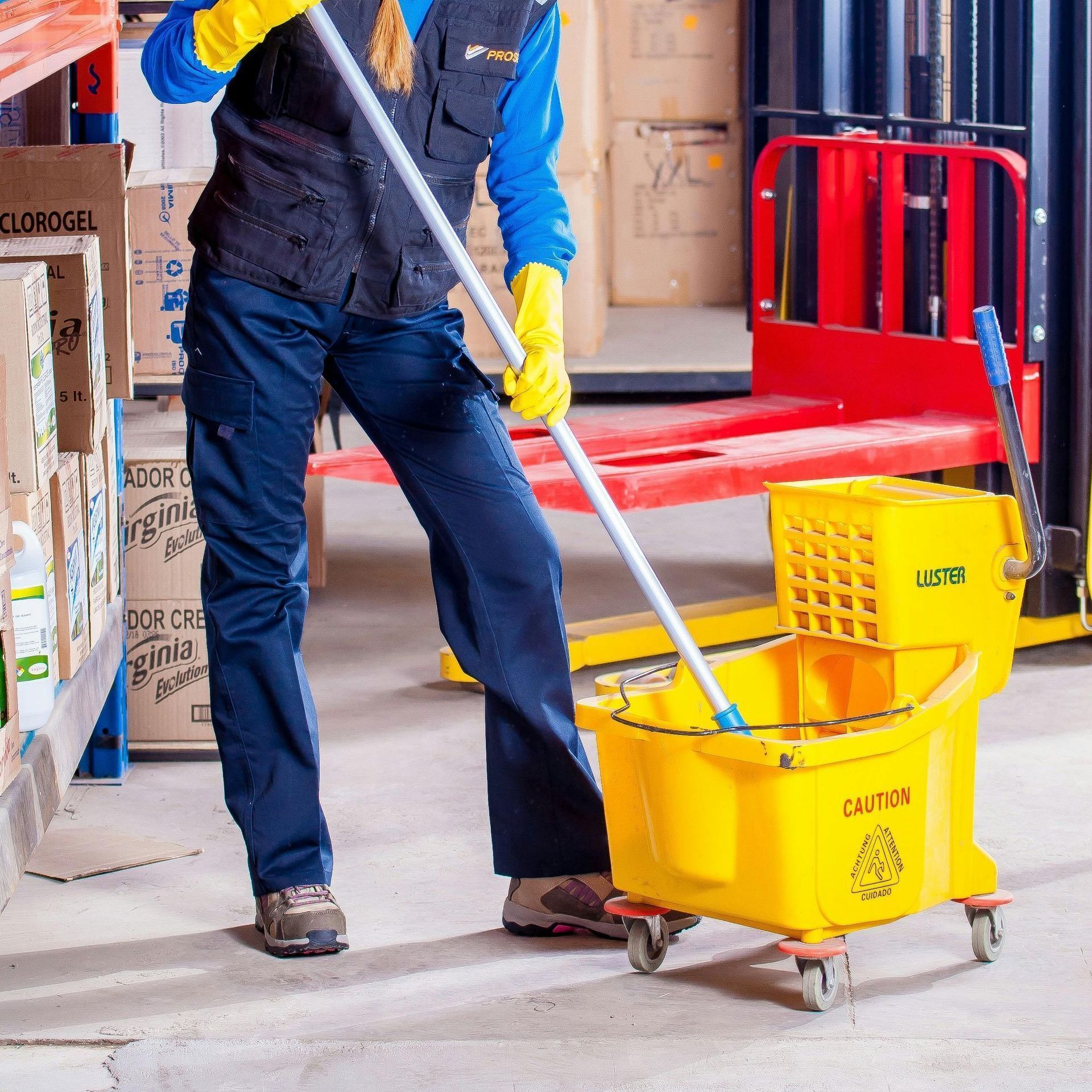 A woman is mopping the floor in a warehouse with a yellow bucket that says caution on it