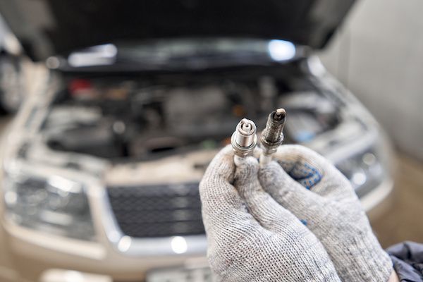 Gloved hands hold a new and used spark plug in front of a car's open hood in a garage. | Dave's Automotive LLC