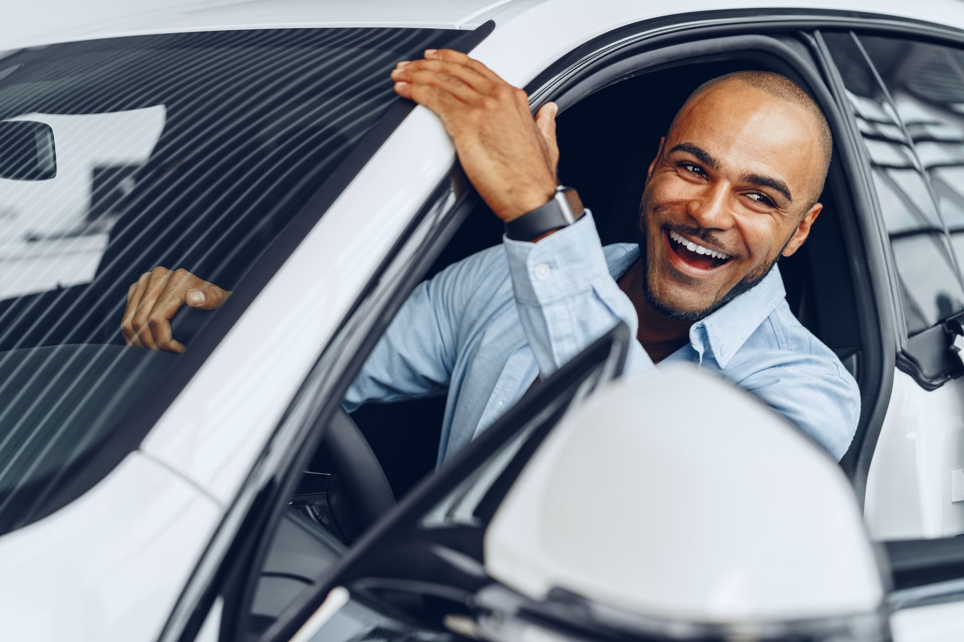 Man smiles, sitting inside a white car, hand on the roof, likely excited about purchase. | Dave's Automotive LLC