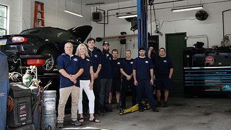 A group of people in blue shirts and khaki pants, standing in a garage. A black car is lifted. | Dave's Automotive LLC