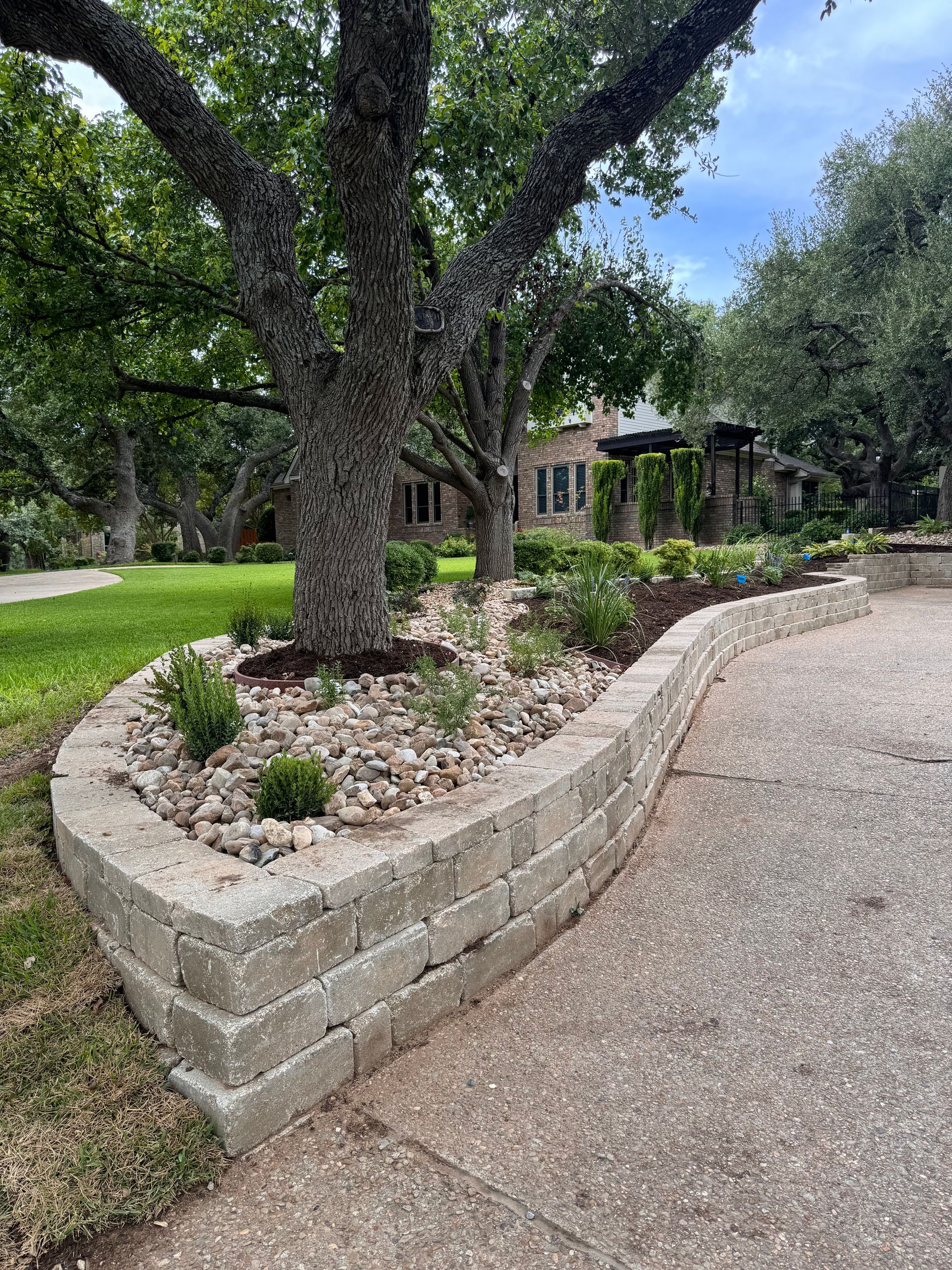 A retaining wall of gray blocks surrounds a tree bed filled with rocks, plants, and mulch next to a driveway.