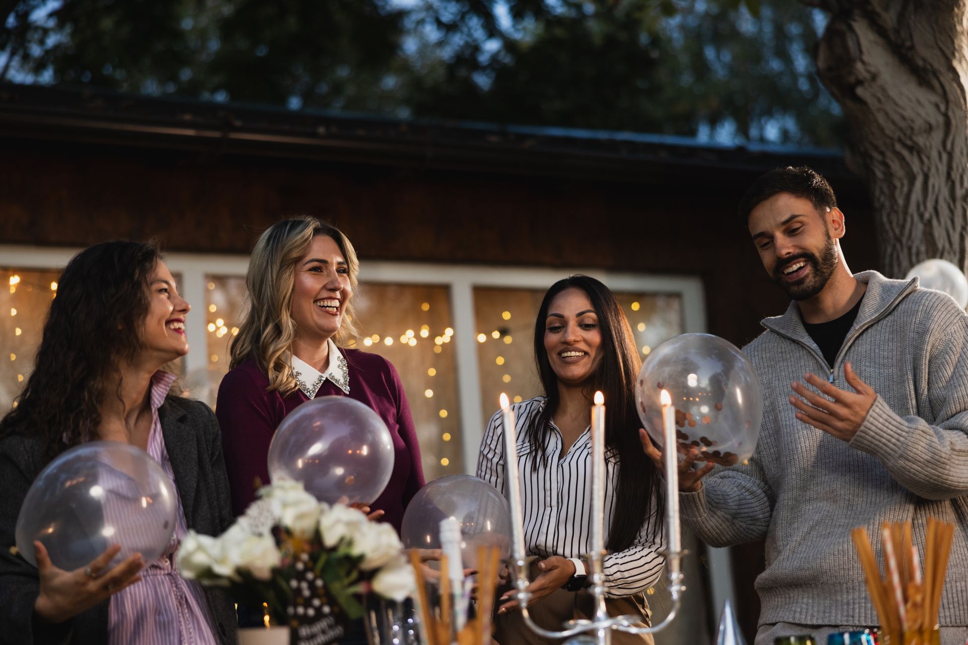 Four smiling people at an outdoor gathering; holding balloons, candles lit; night.