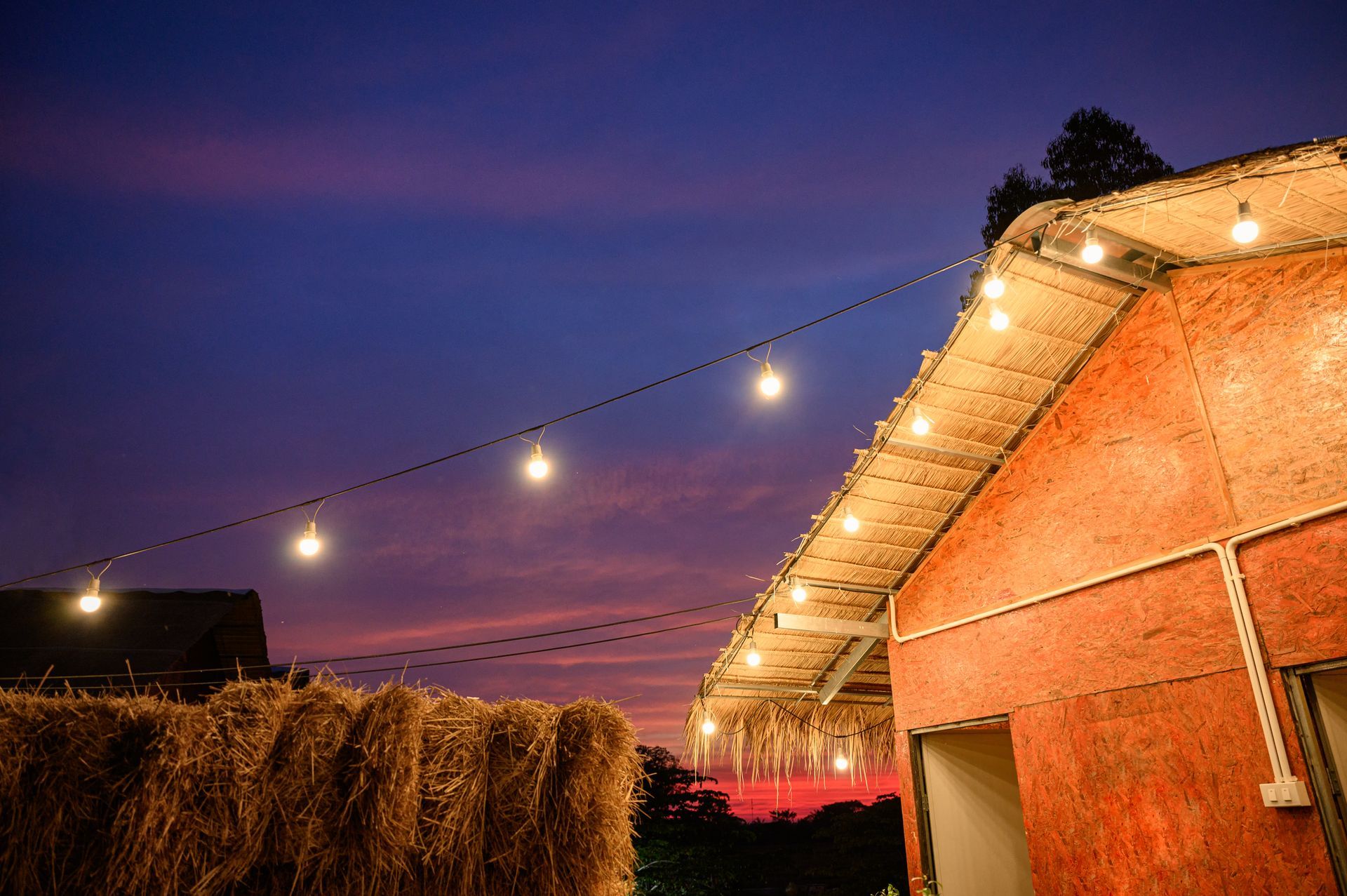 Barn with string lights against a twilight sky, with hay bales in the foreground.
