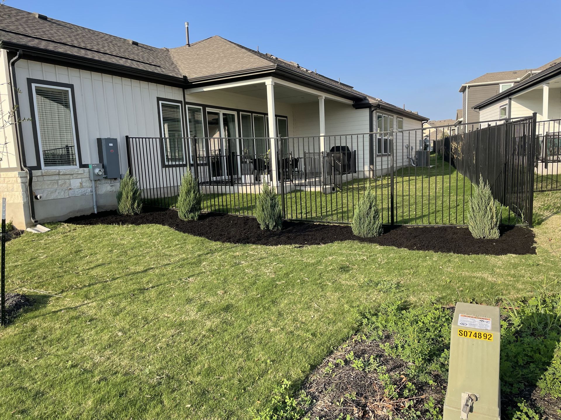 Backyard with a white house, covered patio, black fence, and green grass.