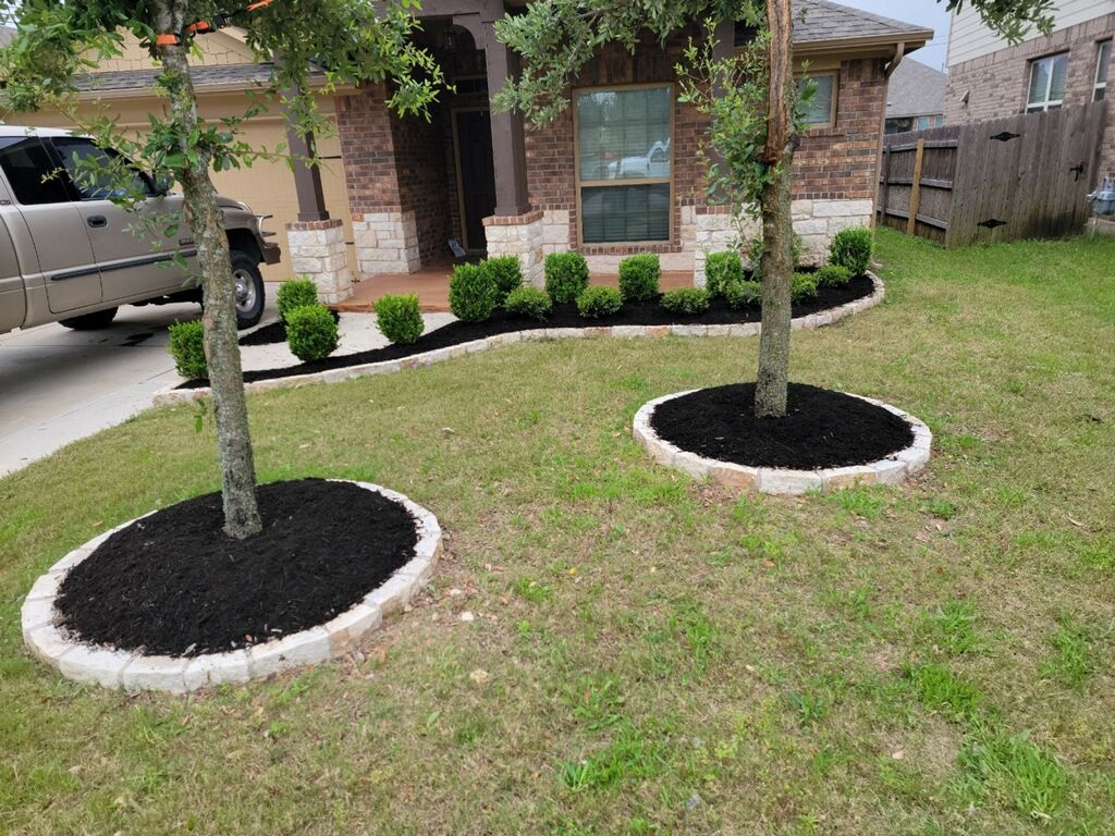 Trees with black mulch beds, bordered by stone, in front of a brick house with manicured landscaping.