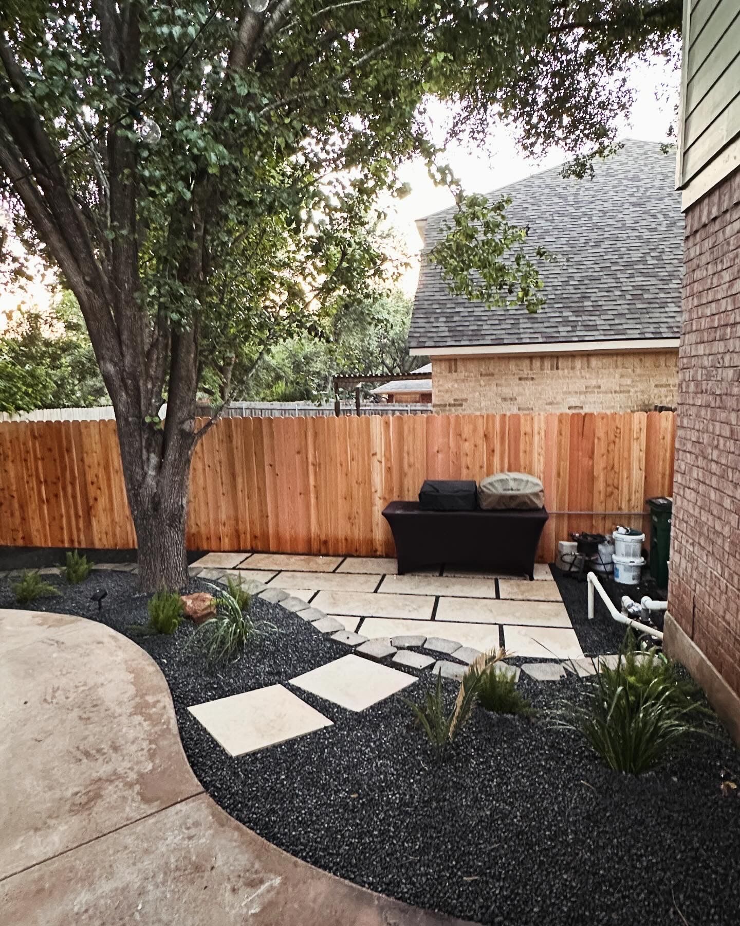 Backyard patio with a wooden fence, a tree, and black rock landscaping.