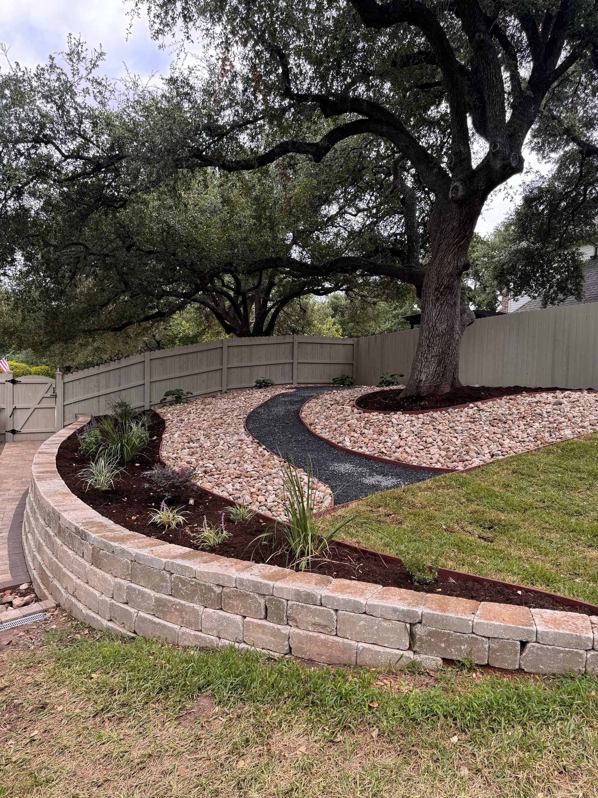 Backyard with a curved retaining wall, rock path, and large tree.