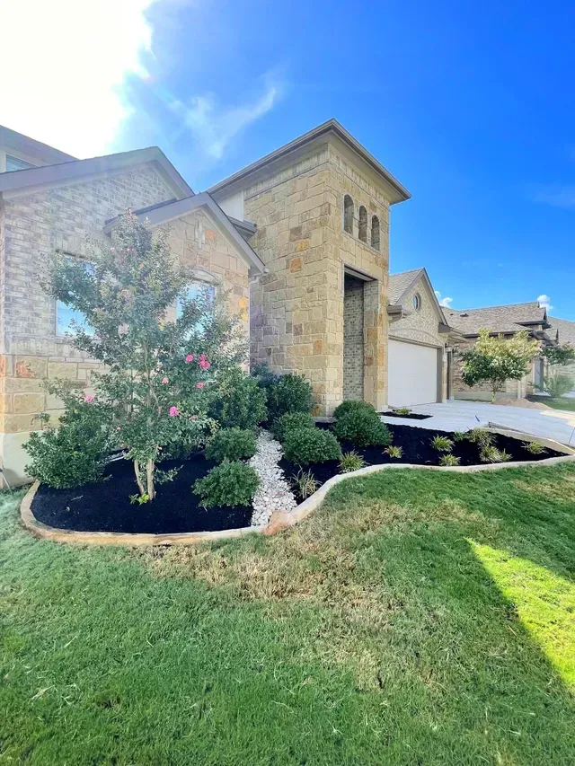 Stone house with landscaped yard, green grass, black mulch, white rocks, and blue sky.