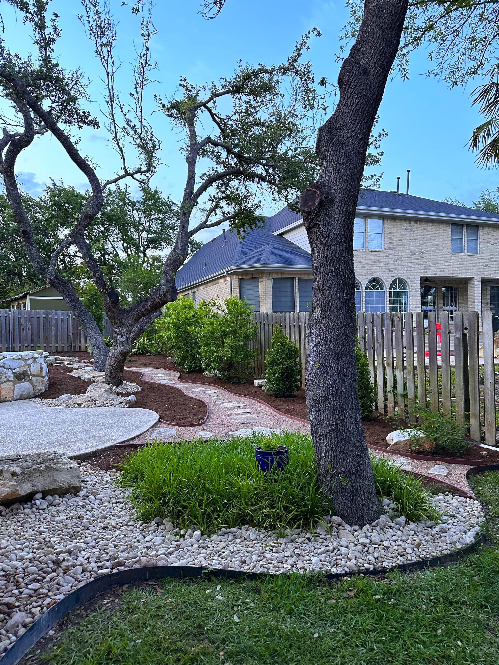 Landscaped yard with gravel, trees, and a brick path leading to a beige brick house with a blue roof.