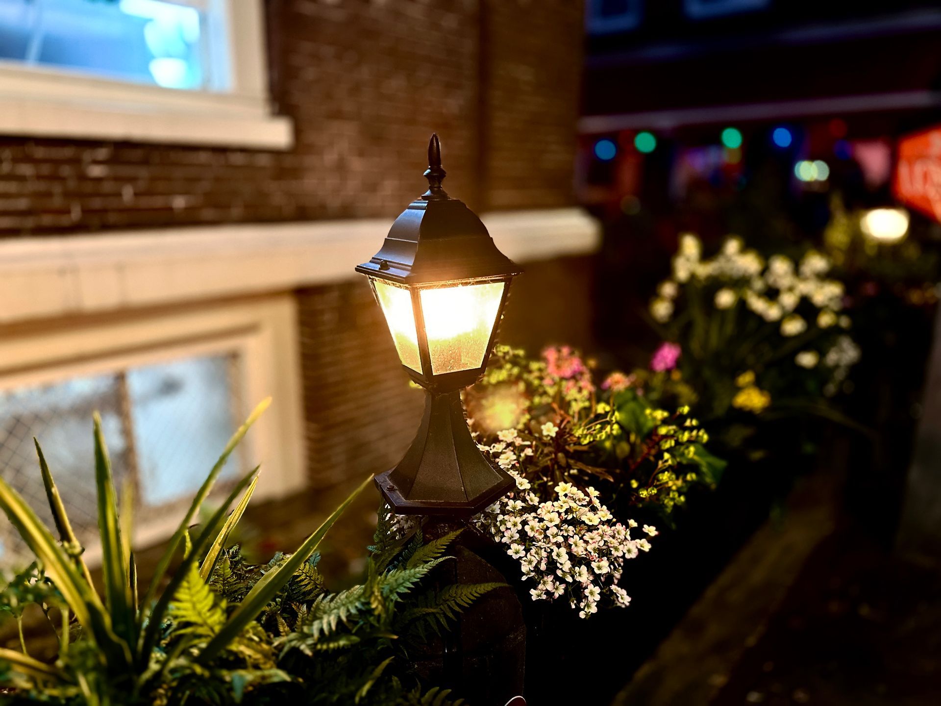 Illuminated lamp post surrounded by plants and flowers, set against a brick wall at night.