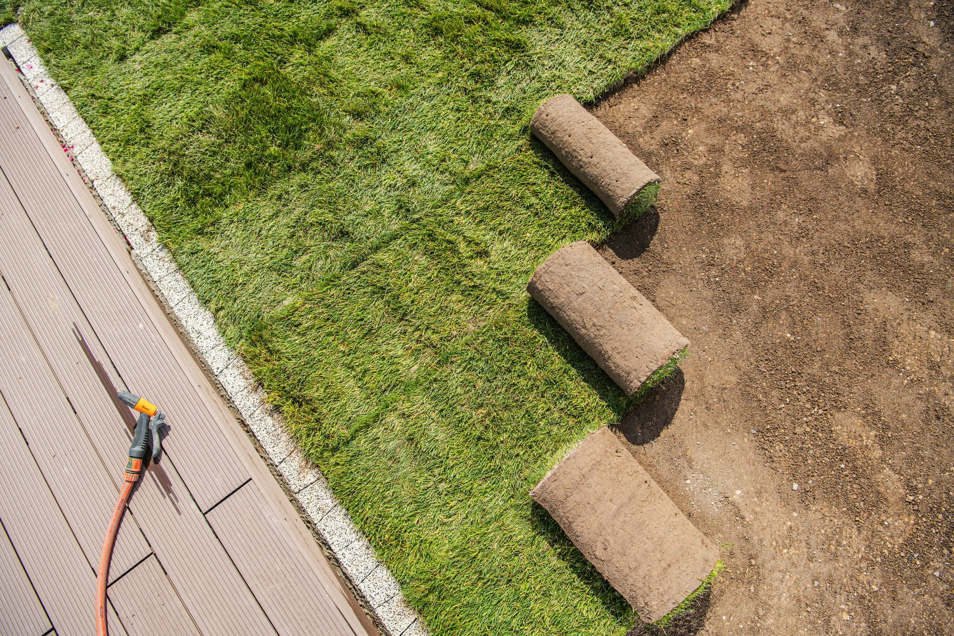 Rolls of sod being laid on dirt next to a patio, with a hose and water nozzle visible.