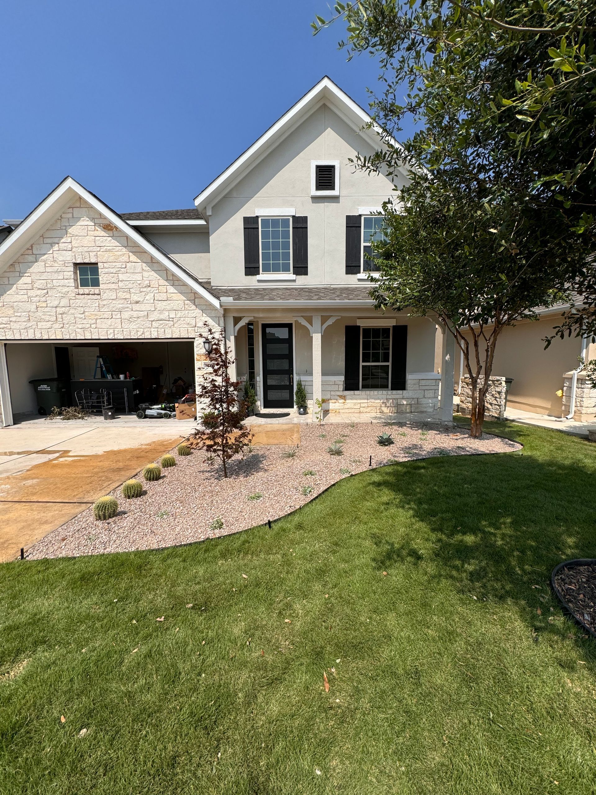 Two-story house with tan stucco and white brick, black shutters, black front door, green lawn, and a sunny sky.