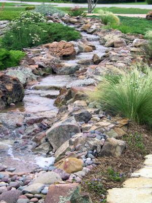 A meandering stream of water flows through a rock garden with lush green plants and grass.