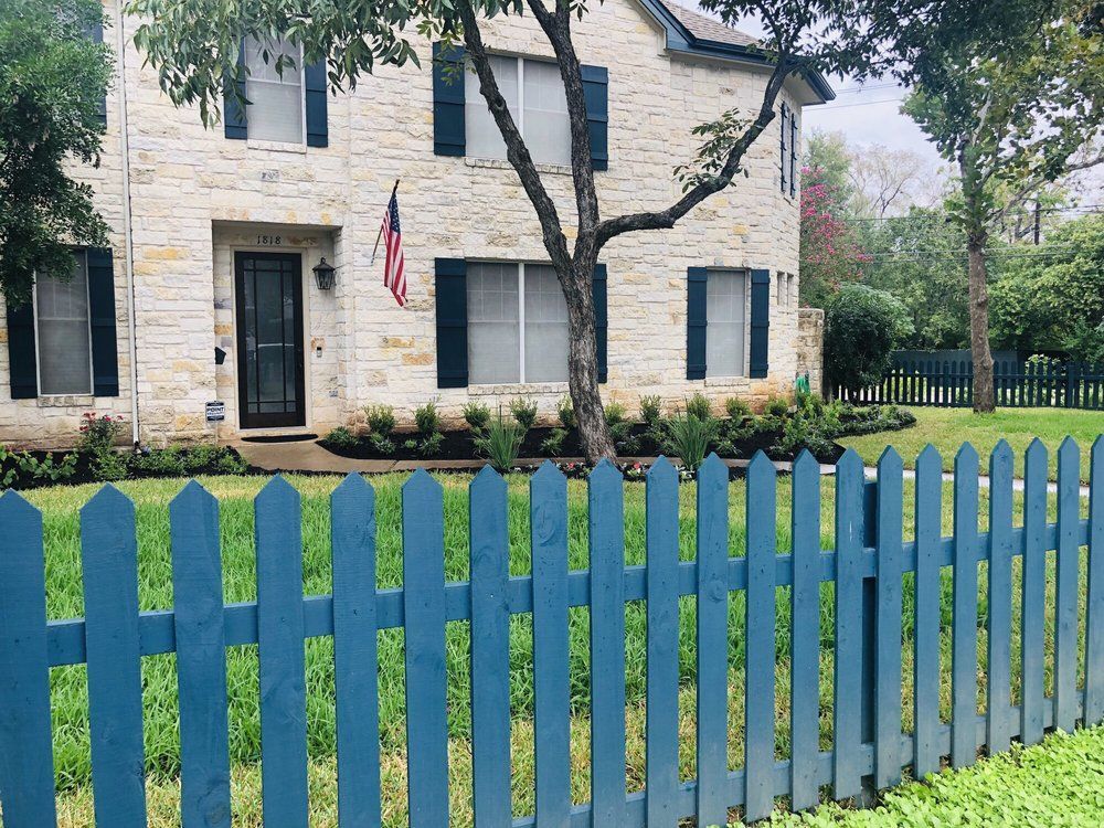 Blue picket fence in front of a two-story stone house with dark shutters; American flag hangs.