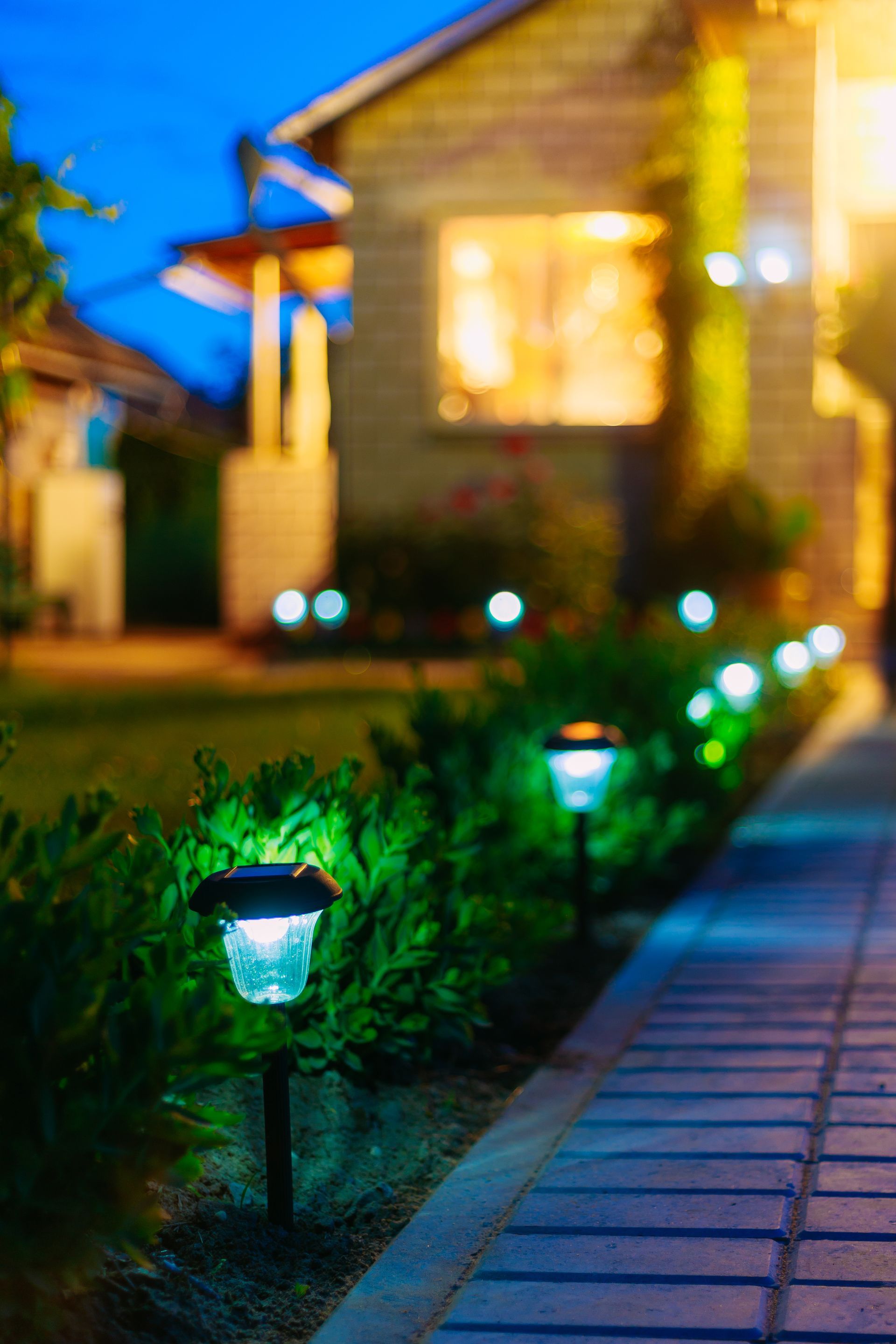 Solar lights illuminate a brick pathway bordering a garden and home at dusk.